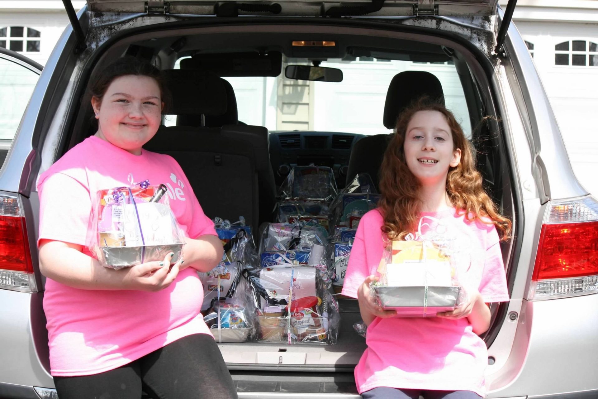 Two girls holding care packages in front of a car's open hatchback, the car is filled with more care packages