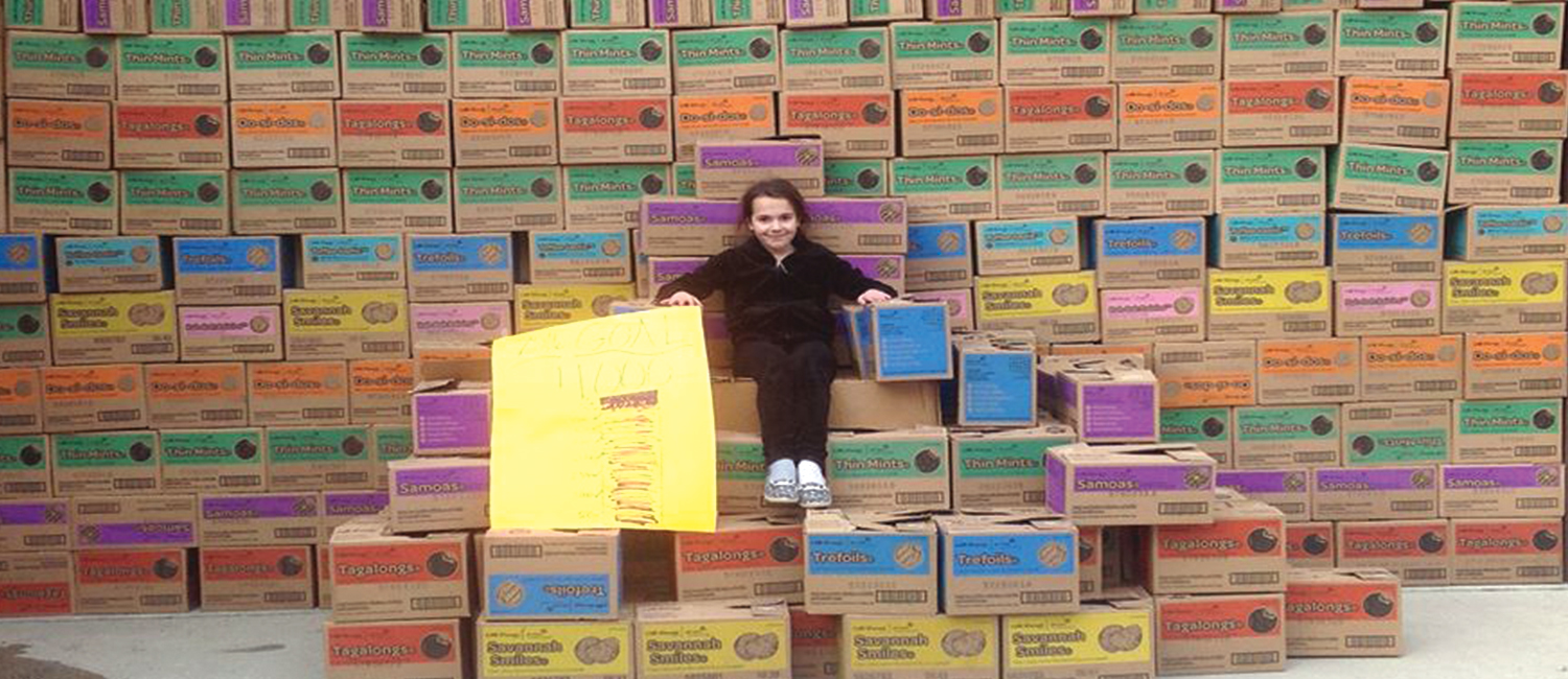  A young girl sitting on a throne made of Girl Scout Cookie cases in front of a wall of cookie cases 