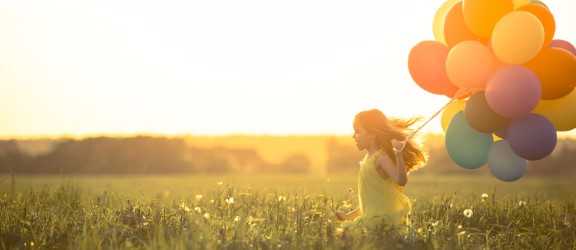 Little girl holding large bunch of ballons running and playing in a field at sunset. 