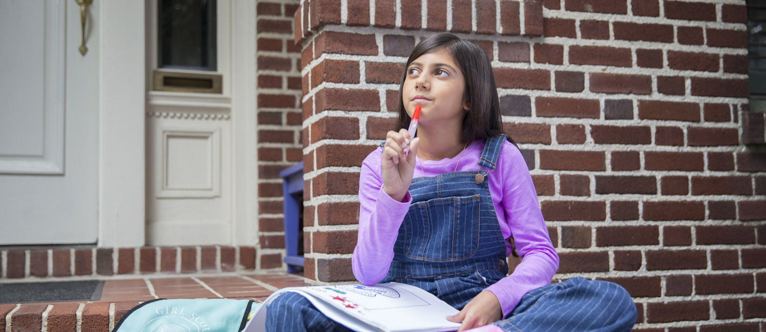 A girl sitting on the front steps of a brick house with a notebook on her lap holding a pen up to her chin with a thoughtful expression 