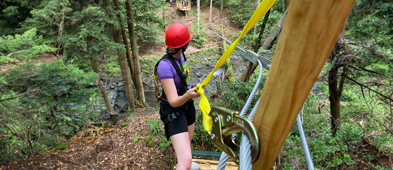 a girl standing on a zip-line platform overlooking a stream and pine trees