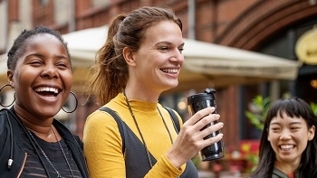 Three girls laughing the one in the middle is holding a reusable to go coffee cup