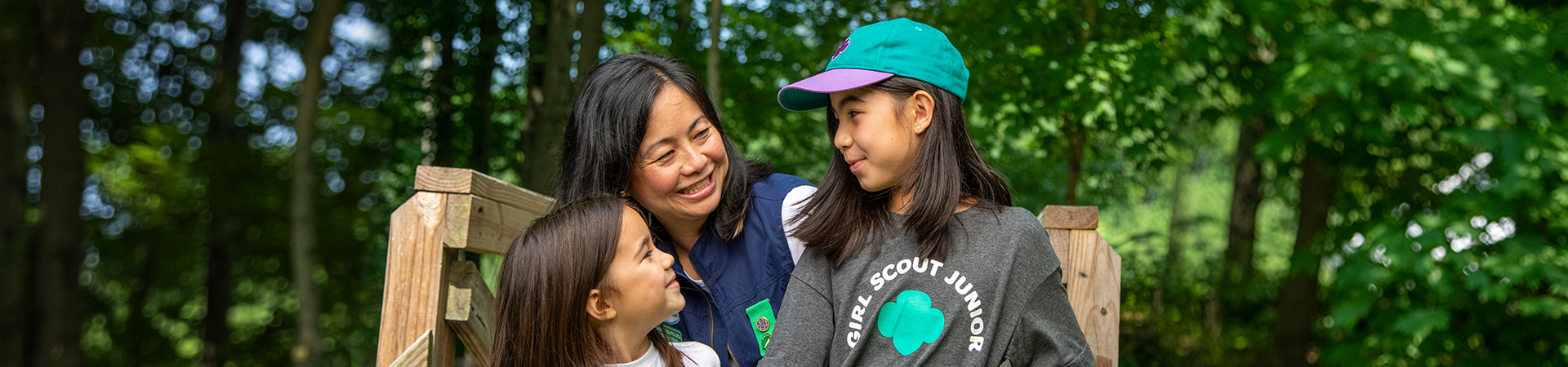  Adult volunteer wearing vest with a girl scout junior (right, in baseball cap) and daisy (left) outside at a park smiling and looking at one another. 
