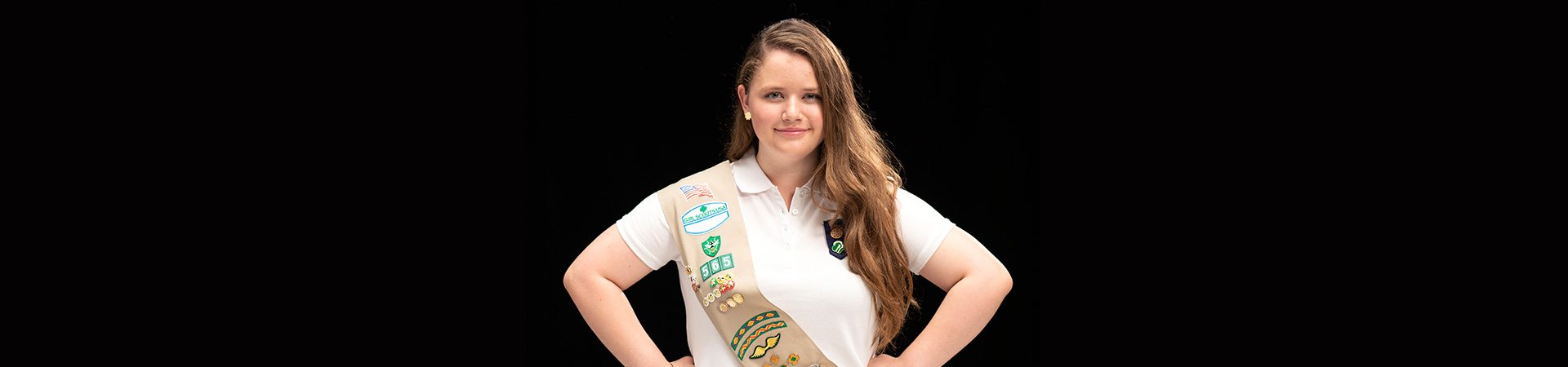  A Gold Award Girl Scout standing in front of a black background with her hands on her hips and a determined smile on her face 