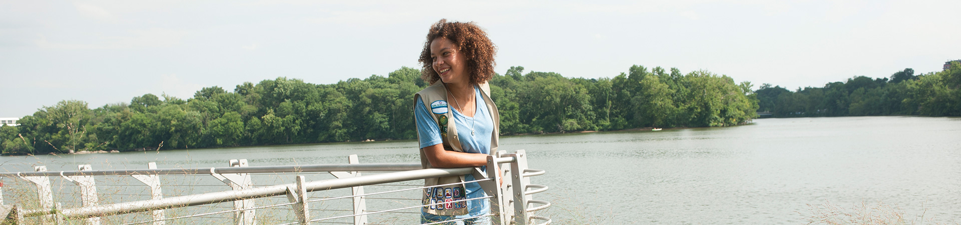  Ambassador Girl Scout standing at a railing in front of a lake looking backwards over her right shoulder and smiling. 