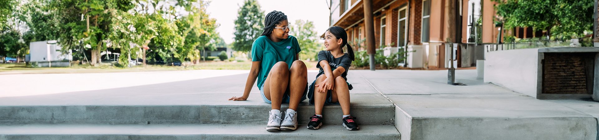  Two girls smiling at each other, sitting on steps outside a school 