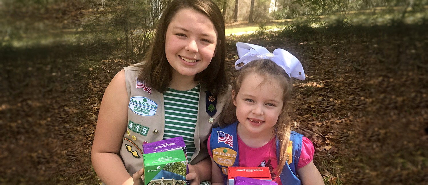  Two girl scouts smiling and holding cookie boxes. 