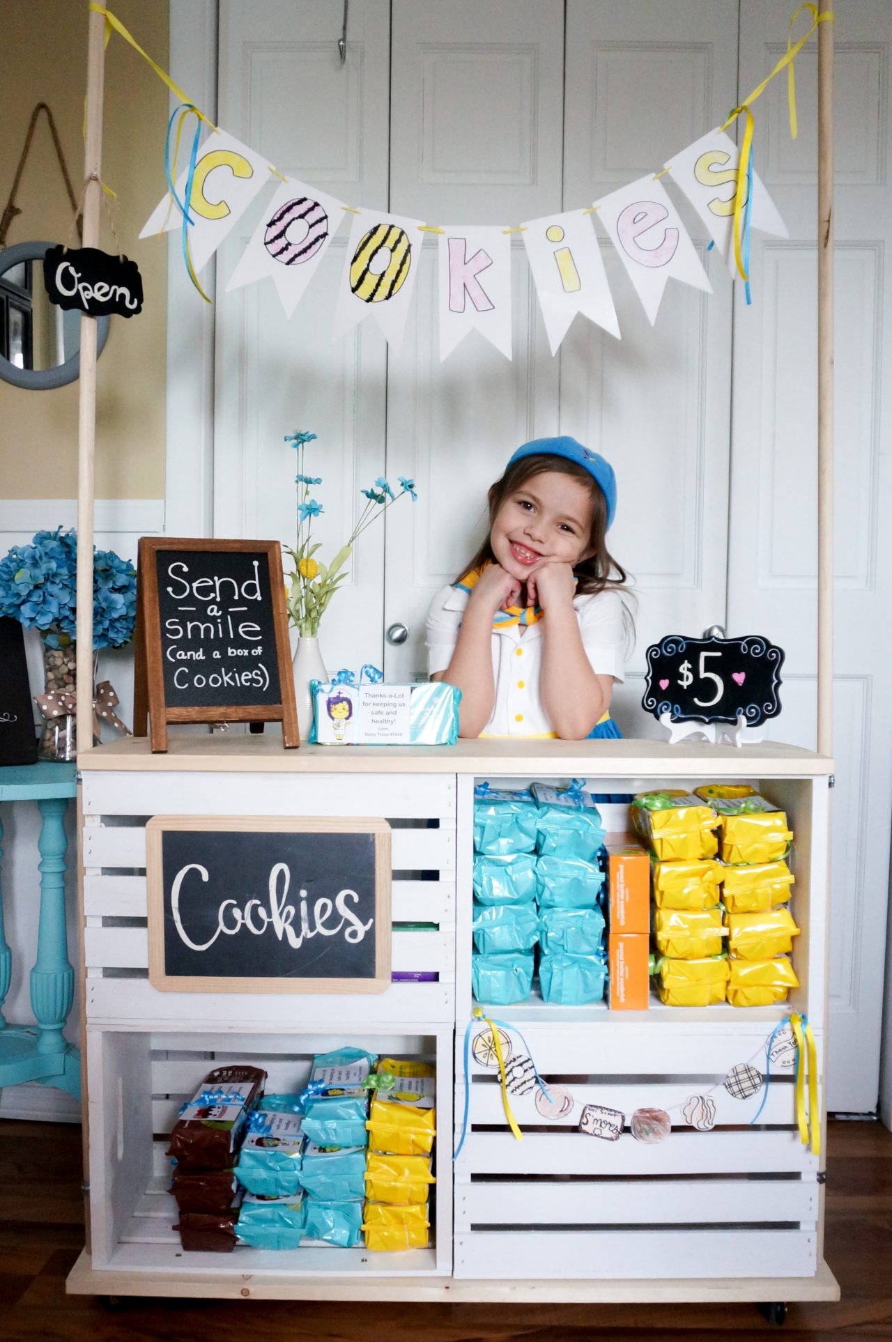 Daisy with her face resting on her fists, standing behind her cookie booth.