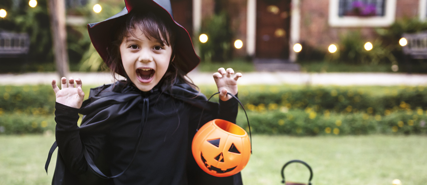  Young girl on Halloween in a witch costume holding a pumpkin-shaped candy bucket trick or treating. 
