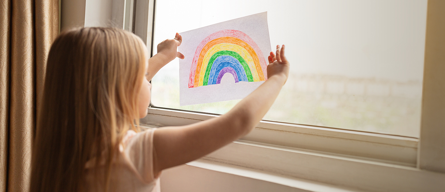  Girl holding painting rainbow against the window during COVID-19 quarantine at home. 