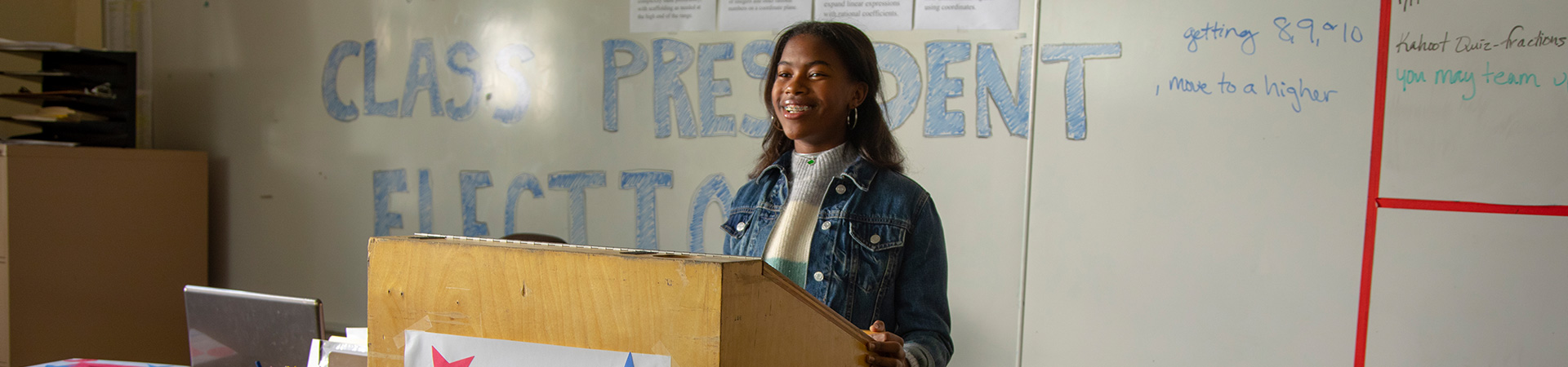  A teen Girl Scout speaking at a lectern in front of a whiteboard that reads "Class President Election"  