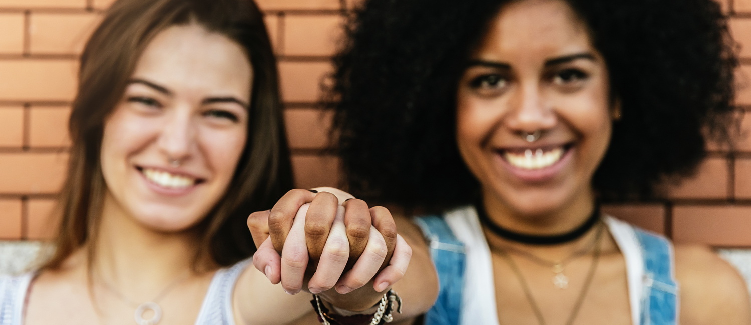  A closeup of two girls' hands with their fingers laced, the girls are smiling in the background in front of a brick wall 